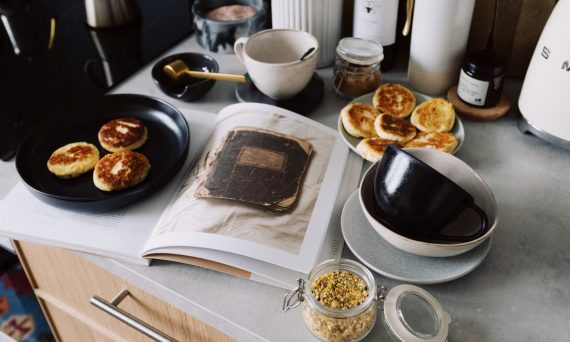 High angle of cookbook and cooked fritters on plates placed on table near utensils and ingredients in cozy kitchen at home